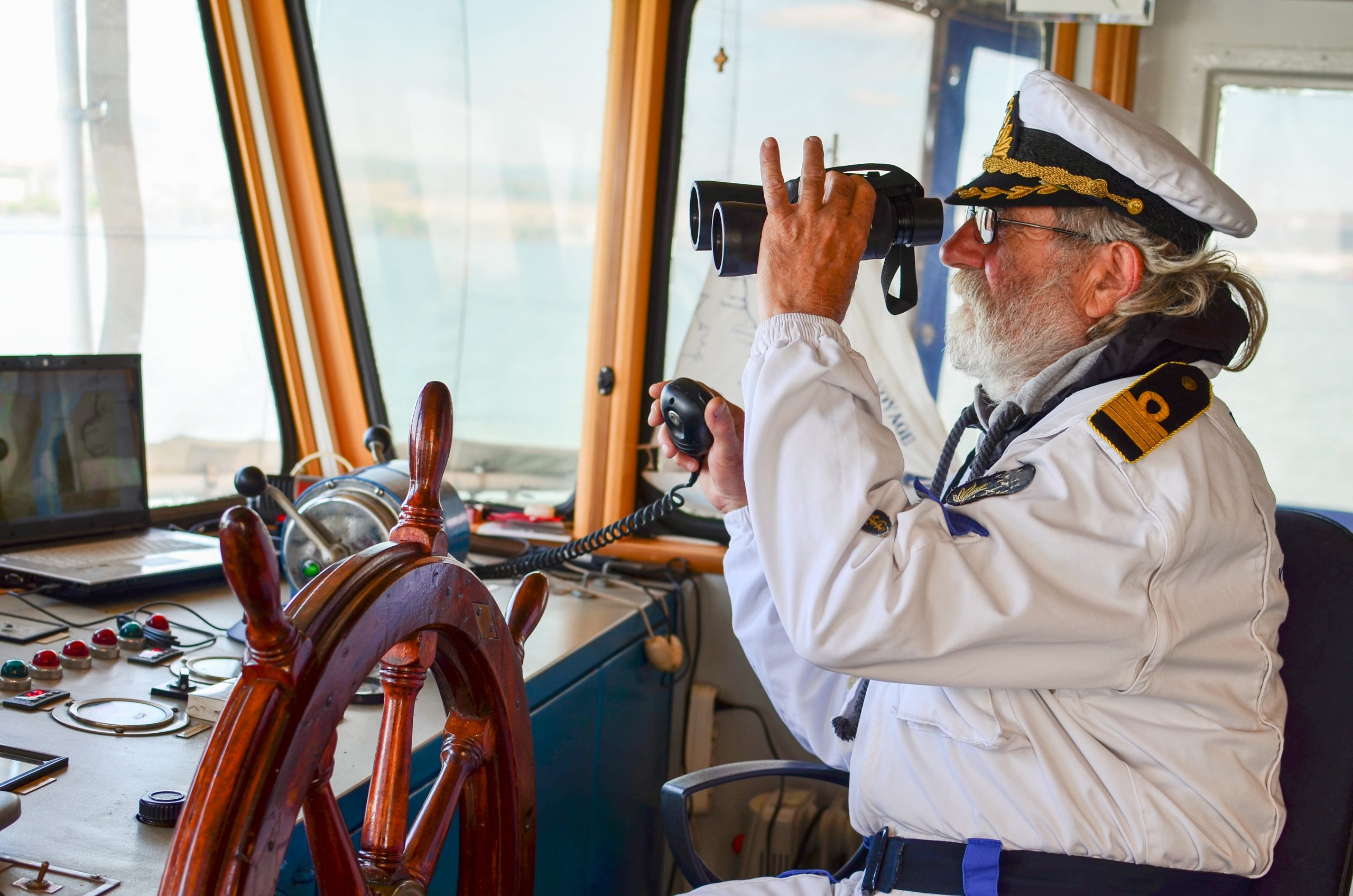 Old experienced captain observes using binoculars by left hand and holding radio comunication equipment by right hand